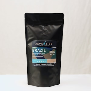 May include: A black bag of coffee beans labeled "JAVA JIVE COFFEE COMPANY" with the words "BRAZIL" and "TIME TO GET YOUR OLE ON!" printed on the front. The bag also includes a roast level indicator and the website address "JAVAJIVEROASTERS.COM".