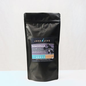May include: A black coffee bag with a label that reads "Java Jive Coffee Company" and "Sumozilla." The label features a graphic of a sumo wrestler. The bag is sealed and has a small circular vent. The background is white.