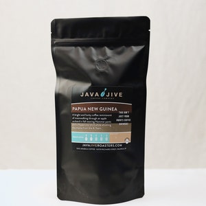 May include: A black coffee bag with a label that reads "PAPUA NEW GUINEA" from Java Jive Coffee Company. The label includes text describing the coffee's flavor profile and roast level. The bag is sealed and has a degassing valve.
