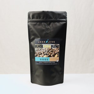 May include: A black coffee bag with a label that reads "JAVA JIVE COFFEE COMPANY BLACK GOLD BLEND." The label features an image of coffee beans and text indicating the roast level. The bag is sealed and has a small air release valve.