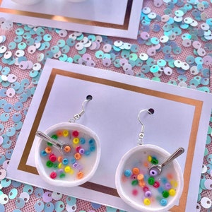 Puede incluir: Un par de pendientes con un cuenco de cerámica blanca lleno de cereales coloridos y una pequeña cuchara de plata. Los pendientes cuelgan de ganchos de plata.