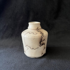 May include: A small, off-white ceramic vase with a rounded body and a narrow neck. The vase features dark, lightning-like patterns across its surface. The handmade pottery has a textured appearance, suggesting a rustic or artisanal style.