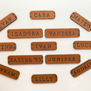 Personalized leather labels with different names or dates written on them, ready to be sewn, are on a white surface.
