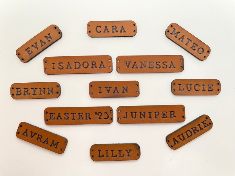 Personalized leather labels with different names or dates written on them, ready to be sewn, are on a white surface.