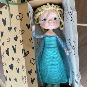 Handmade crocheted amigurumi dolls of Elsa and Anna, inspired by Frozen. Detailed with colorful outfits, perfect for custom name personalization or gifting. Lying inside a gift box.