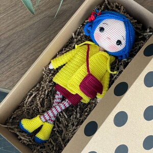 A handmade amigurumi Coraline crochet doll with blue hair, yellow raincoat, striped leggings, yellow boots, and a red shoulder bag. Personalized name tag option available. Perfect handmade gift for kids, collectors, and Coraline movie fans.
