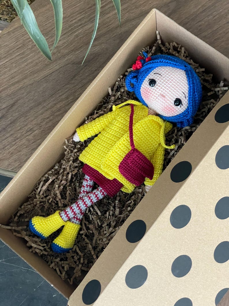A handmade amigurumi Coraline crochet doll with blue hair, yellow raincoat, striped leggings, yellow boots, and a red shoulder bag. Personalized name tag option available. Perfect handmade gift for kids, collectors, and Coraline movie fans.