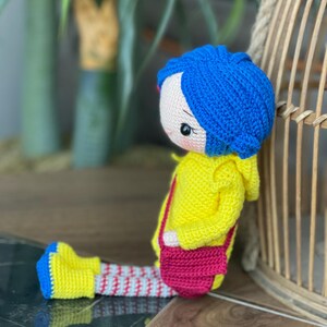 A handmade amigurumi Coraline crochet doll with blue hair, yellow raincoat, striped leggings, yellow boots, and a red shoulder bag. Personalized name tag option available. Perfect handmade gift for kids, collectors, and Coraline movie fans.