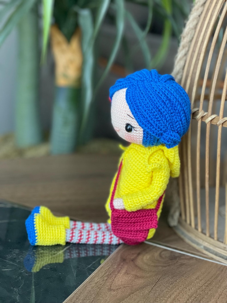 A handmade amigurumi Coraline crochet doll with blue hair, yellow raincoat, striped leggings, yellow boots, and a red shoulder bag. Personalized name tag option available. Perfect handmade gift for kids, collectors, and Coraline movie fans.