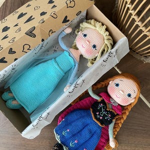 Handmade crocheted amigurumi dolls of Elsa and Anna, inspired by Frozen. Detailed with colorful outfits, perfect for custom name personalization or gifting. Lying inside a gift box.
