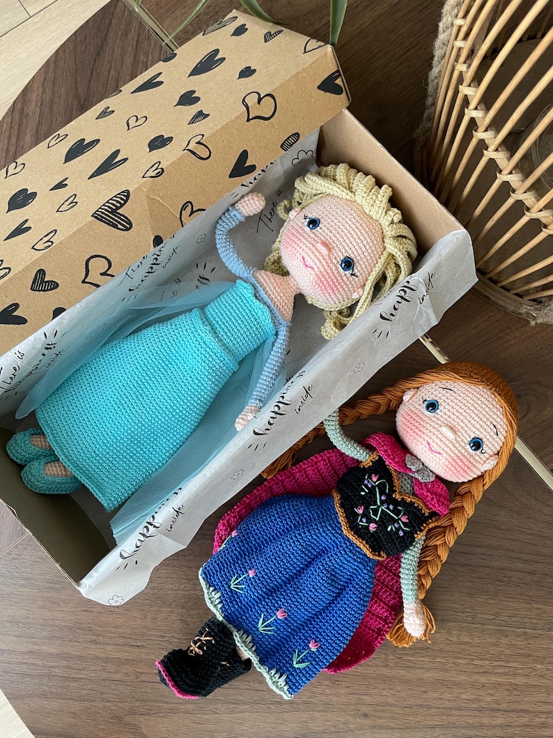 Handmade crocheted amigurumi dolls of Elsa and Anna, inspired by Frozen. Detailed with colorful outfits, perfect for custom name personalization or gifting. Lying inside a gift box.