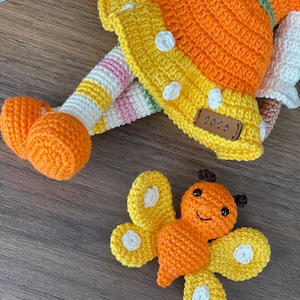 Handmade crocheted amigurumi doll named Orange Girl, featuring a personalized leather name label and accompanied by a marmalade-colored butterfly pet. Perfect gift for children and collectors.