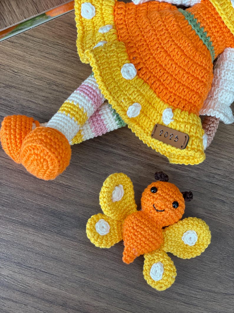 Handmade crocheted amigurumi doll named Orange Girl, featuring a personalized leather name label and accompanied by a marmalade-colored butterfly pet. Perfect gift for children and collectors.