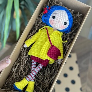 A handmade amigurumi Coraline crochet doll with blue hair, yellow raincoat, striped leggings, yellow boots, and a red shoulder bag. Personalized name tag option available. Perfect handmade gift for kids, collectors, and Coraline movie fans.