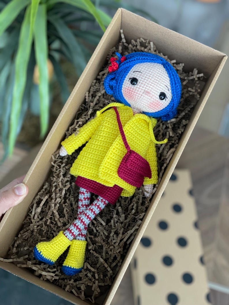 A handmade amigurumi Coraline crochet doll with blue hair, yellow raincoat, striped leggings, yellow boots, and a red shoulder bag. Personalized name tag option available. Perfect handmade gift for kids, collectors, and Coraline movie fans.