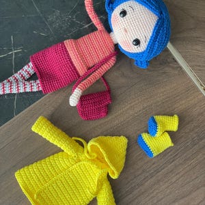 A handmade amigurumi Coraline crochet doll with blue hair, yellow raincoat, striped leggings, yellow boots, and a red shoulder bag. Personalized name tag option available. Perfect handmade gift for kids, collectors, and Coraline movie fans.