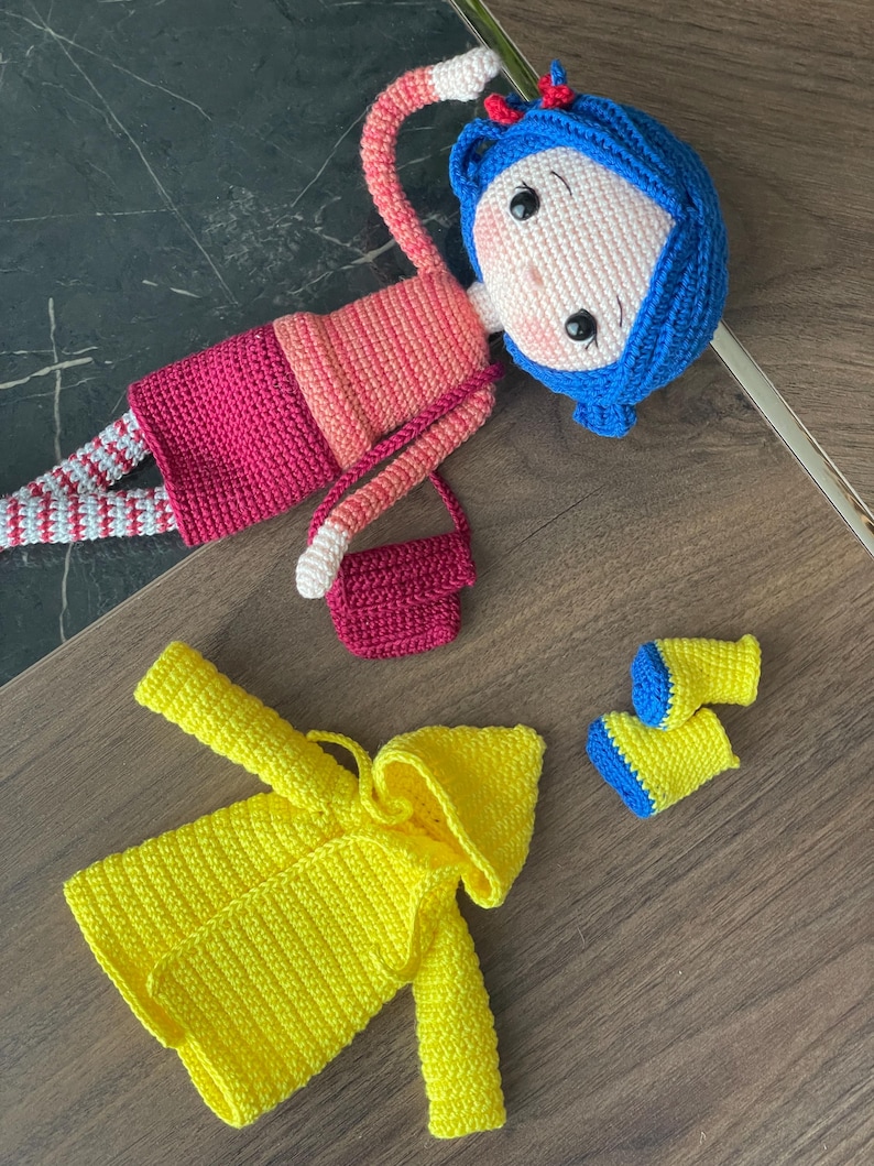 A handmade amigurumi Coraline crochet doll with blue hair, yellow raincoat, striped leggings, yellow boots, and a red shoulder bag. Personalized name tag option available. Perfect handmade gift for kids, collectors, and Coraline movie fans.
