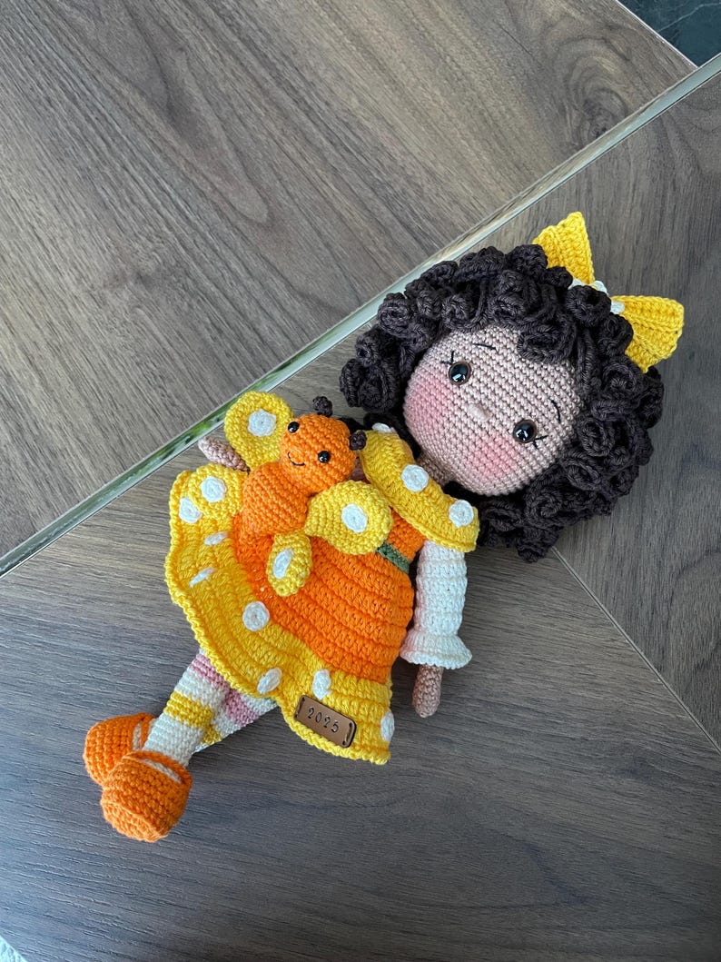 Handmade crocheted amigurumi doll named Orange Girl, featuring a personalized leather name label and accompanied by a marmalade-colored butterfly pet. Perfect gift for children and collectors.