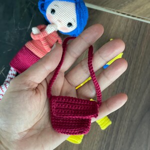 A handmade amigurumi Coraline crochet doll with blue hair, yellow raincoat, striped leggings, yellow boots, and a red shoulder bag. Personalized name tag option available. Perfect handmade gift for kids, collectors, and Coraline movie fans.