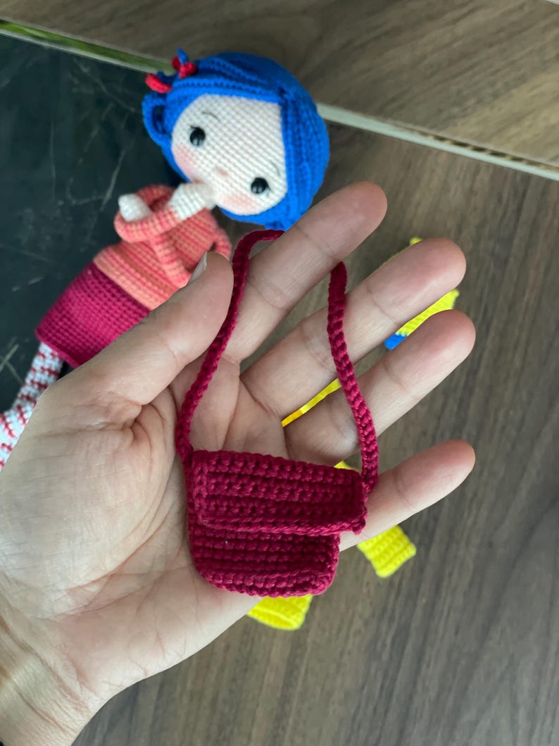 A handmade amigurumi Coraline crochet doll with blue hair, yellow raincoat, striped leggings, yellow boots, and a red shoulder bag. Personalized name tag option available. Perfect handmade gift for kids, collectors, and Coraline movie fans.