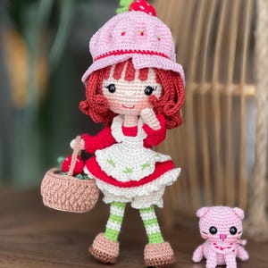 Könnte beinhalten: Handgefertigte Häkelpuppe, die ein Erdbeermädchen darstellt, mit einem rosa Hut mit Erdbeere, rotem Haar und einem weißen Kleid mit rotem Oberteil. Sie hält einen Korb mit Erdbeeren. Eine kleine rosa Häkelkatze ist daneben.