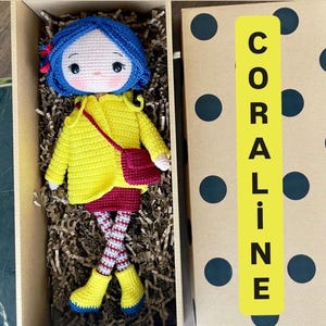 A handmade amigurumi Coraline crochet doll with blue hair, yellow raincoat, striped leggings, yellow boots, and a red shoulder bag. Personalized name tag option available. Perfect handmade gift for kids, collectors, and Coraline movie fans.