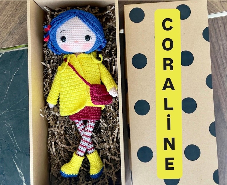 A handmade amigurumi Coraline crochet doll with blue hair, yellow raincoat, striped leggings, yellow boots, and a red shoulder bag. Personalized name tag option available. Perfect handmade gift for kids, collectors, and Coraline movie fans.
