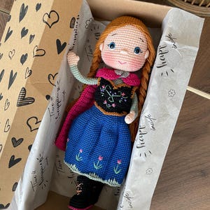 Handmade crocheted amigurumi doll of Anna, inspired by Frozen. Detailed with colorful outfits, perfect for custom name personalization or gifting. Lying inside a gift box.