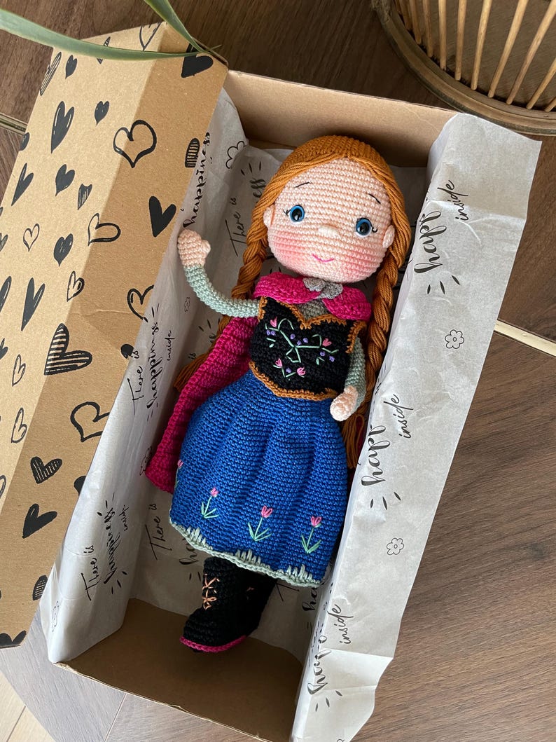 Handmade crocheted amigurumi doll of Anna, inspired by Frozen. Detailed with colorful outfits, perfect for custom name personalization or gifting. Lying inside a gift box.