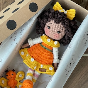 Handmade crocheted amigurumi doll named Orange Girl, featuring a personalized leather name label and accompanied by a marmalade-colored butterfly pet. Perfect gift for children and collectors. Lying inside a gift box.