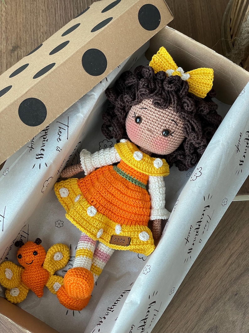 Handmade crocheted amigurumi doll named Orange Girl, featuring a personalized leather name label and accompanied by a marmalade-colored butterfly pet. Perfect gift for children and collectors. Lying inside a gift box.