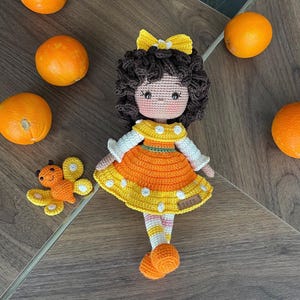 Handmade crocheted amigurumi doll named Orange Girl, featuring a personalized leather name label and accompanied by a marmalade-colored butterfly pet. Perfect gift for children and collectors.