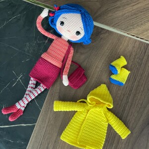 A handmade amigurumi Coraline crochet doll with blue hair, yellow raincoat, striped leggings, yellow boots, and a red shoulder bag. Personalized name tag option available. Perfect handmade gift for kids, collectors, and Coraline movie fans.