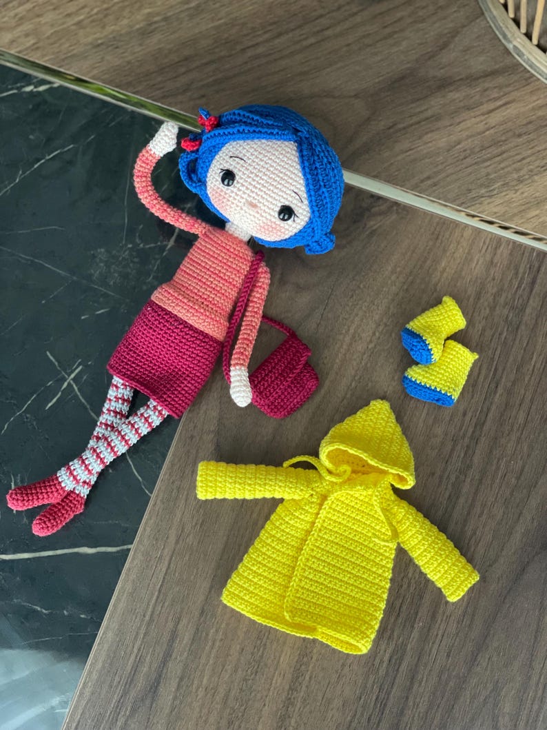 A handmade amigurumi Coraline crochet doll with blue hair, yellow raincoat, striped leggings, yellow boots, and a red shoulder bag. Personalized name tag option available. Perfect handmade gift for kids, collectors, and Coraline movie fans.