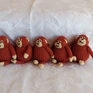 May include: Five knitted monkey toys in a row. The monkeys are brown with cream-colored faces and limbs. They are sitting on a white fabric surface.