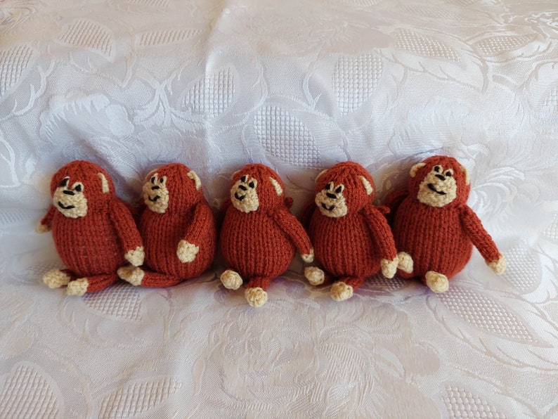 May include: Five knitted monkey toys in a row. The monkeys are brown with cream-colored faces and limbs. They are sitting on a white fabric surface.