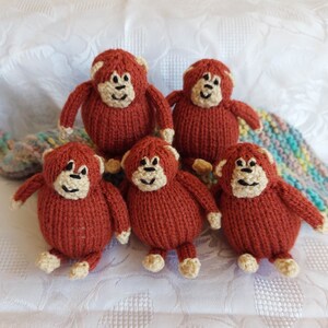 May include: Five knitted brown monkey toys with beige faces and black eyes. The monkeys are sitting on a white surface.