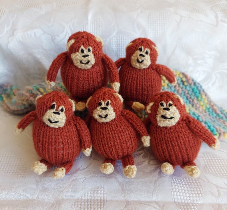 May include: Five knitted brown monkey toys with beige faces and black eyes. The monkeys are sitting on a white surface.