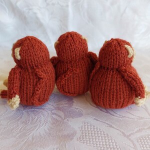 May include: Three knitted monkey toys in a reddish-brown color with cream-colored accents. The monkeys are sitting in a row, facing away from the camera.