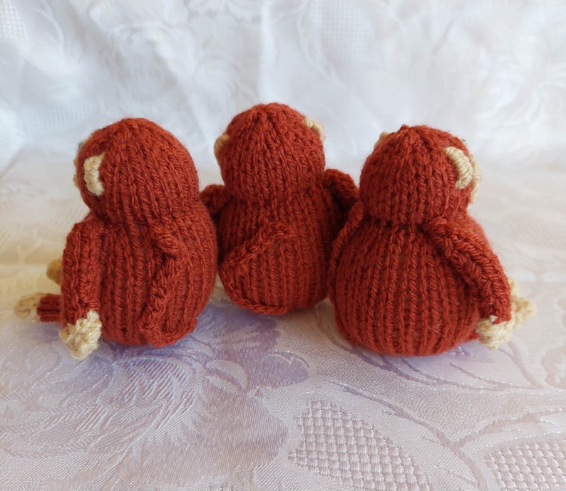 May include: Three knitted monkey toys in a reddish-brown color with cream-colored accents. The monkeys are sitting in a row, facing away from the camera.