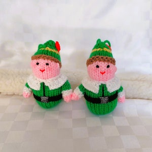May include: Two knitted elf dolls. Each doll has a pink face, a green outfit with a white collar, and a green hat with gold trim. The elves are holding hands. One elf has a red flower on its hat.