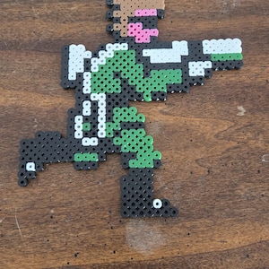 May include: A pixel art figure made of colorful plastic beads. The figure is in a running pose, holding a weapon. The color scheme includes black, green, white, brown, and pink. The figure is on a wooden surface.