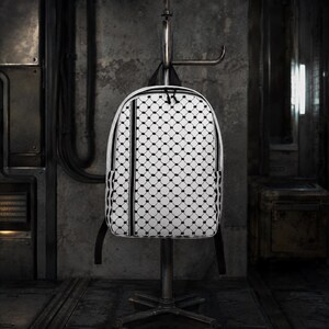 Könnte beinhalten: Weißer Rucksack mit einem schwarzen geometrischen Muster. Der Rucksack hat schwarze Träger und einen schwarzen Reißverschluss. Das Design zeigt ein sich wiederholendes Muster aus schwarzen Punkten in einem Raster auf weißem Hintergrund. Der Rucksack hängt an einem Metallständer.