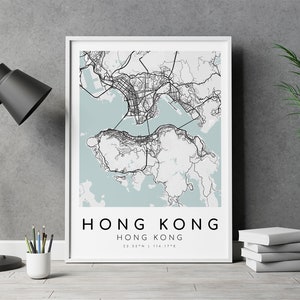 May include: A framed map print of Hong Kong, featuring a detailed black and white cartographic design set against a light blue background. The print includes the city's name and coordinates. A black desk lamp, pencils, and a potted plant are also visible.