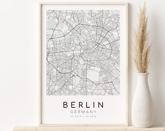 May include: A black and white map print of Berlin, Germany, in a white frame on a wooden shelf. The map shows the city streets and landmarks. The text "BERLIN GERMANY 52.52°N | 13.40°E" is printed below the map.