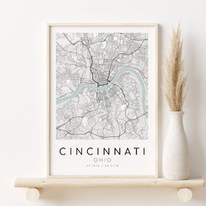 May include: A black and white map print of Cincinnati, Ohio, in a light wood frame. The map shows the city's streets and the Ohio River. The print is on a white background and has the city's name and coordinates at the bottom.