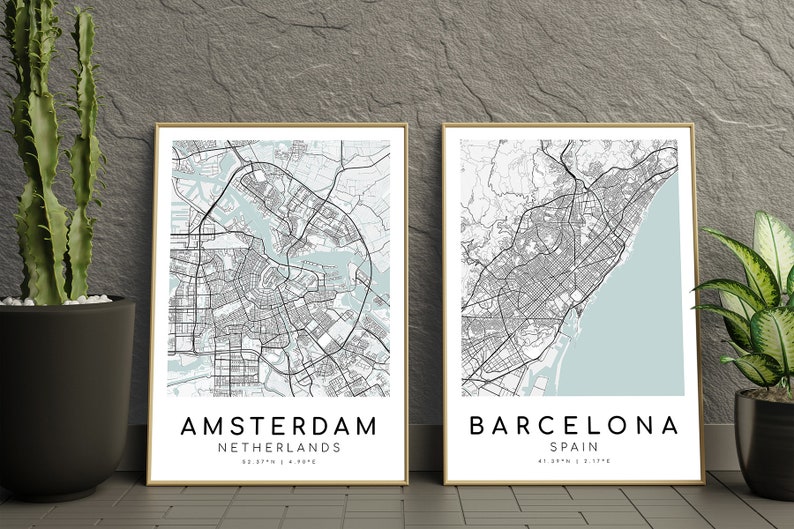 May include: Two framed city map prints. One features Amsterdam, Netherlands, and the other Barcelona, Spain. Both maps are in black and white with a light blue accent for the water areas. The city names and coordinates are printed in a clean, modern font.