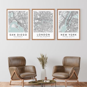 May include: Three framed city map prints. The prints feature San Diego, London, and New York, with light blue water areas and black street details. The frames are a light brown color. The prints are displayed above two brown leather armchairs.