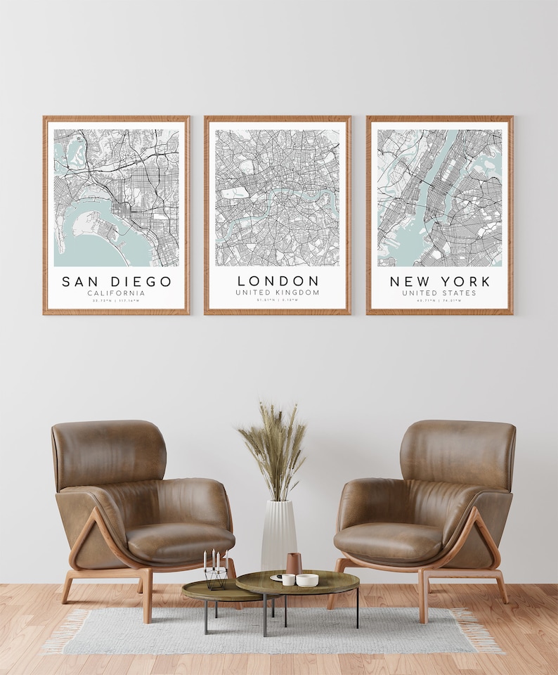 May include: Three framed city map prints. The prints feature San Diego, London, and New York, with light blue water areas and black street details. The frames are a light brown color. The prints are displayed above two brown leather armchairs.