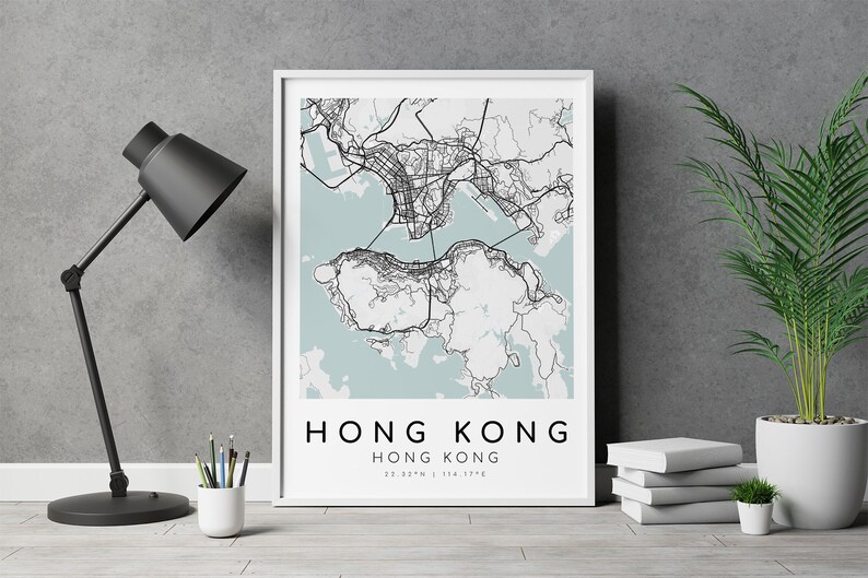 May include: A framed map print of Hong Kong, featuring a detailed black and white cartographic design set against a light blue background. The print includes the city's name and coordinates. A black desk lamp, pencils, and a potted plant are also visible.